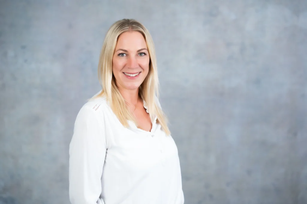 Eine lächelnde Frau mit blonder, glatter Frisur trägt eine weiße Bluse vor einem graublauen Hintergrund.